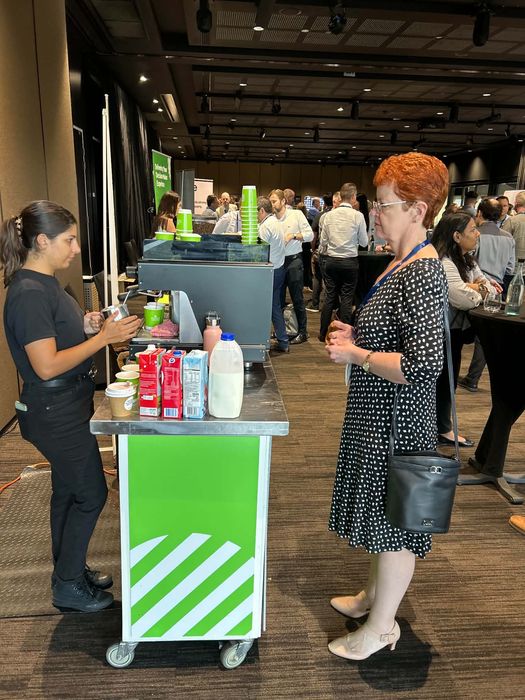 Barista serving coffee at a busy corporate conference — green branded cart with full room of professional attendees